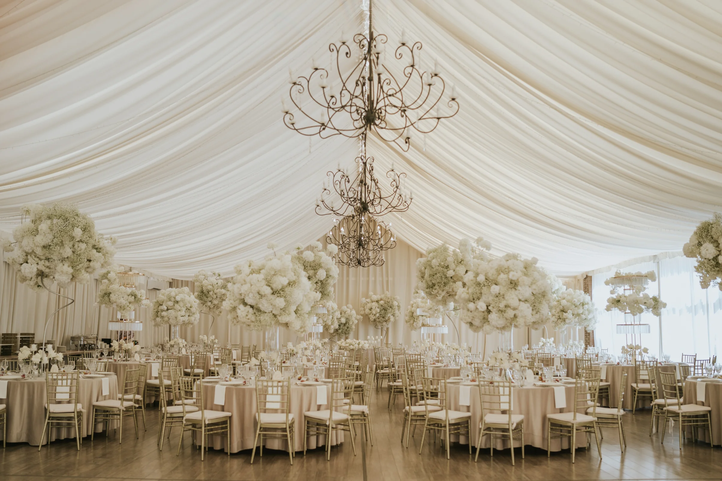 Elegancka sala weselna z białymi obrusami, złotymi krzesłami, kwiatowymi dekoracjami i kryształowymi żyrandolami pod namiotem.
