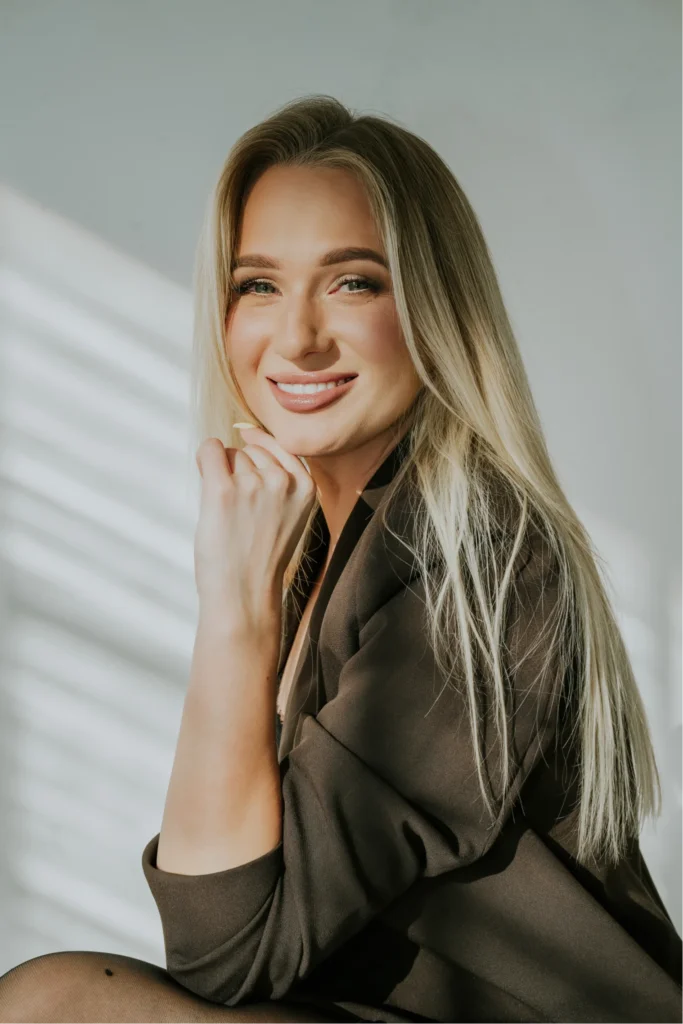 Uśmiechnięta kobieta z długimi blond włosami w oliwkowej marynarce przy białej ścianie z cieniami z okna