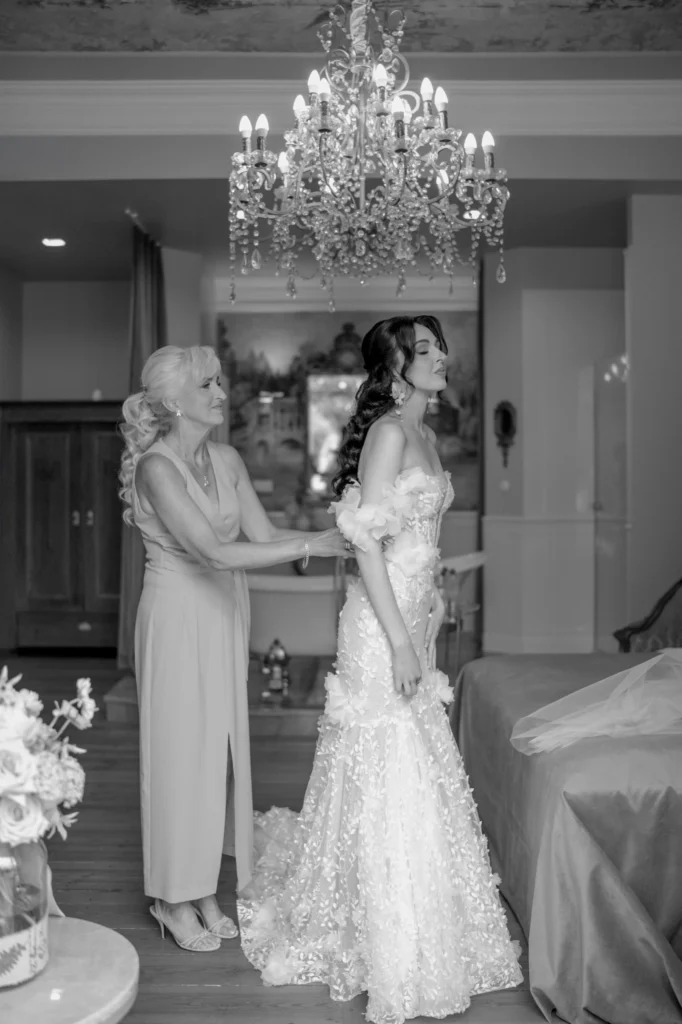 Matka zapina suknię ślubną córce pod kryształowym żyrandolem w eleganckim wnętrzu