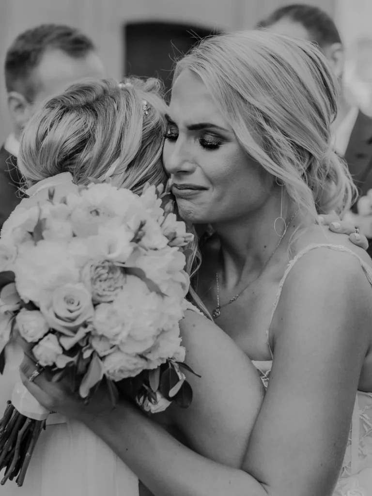 Panna młoda w białej sukni ślubnej przytula się do gościa weselnego trzymając bukiet białych róż i piwonii