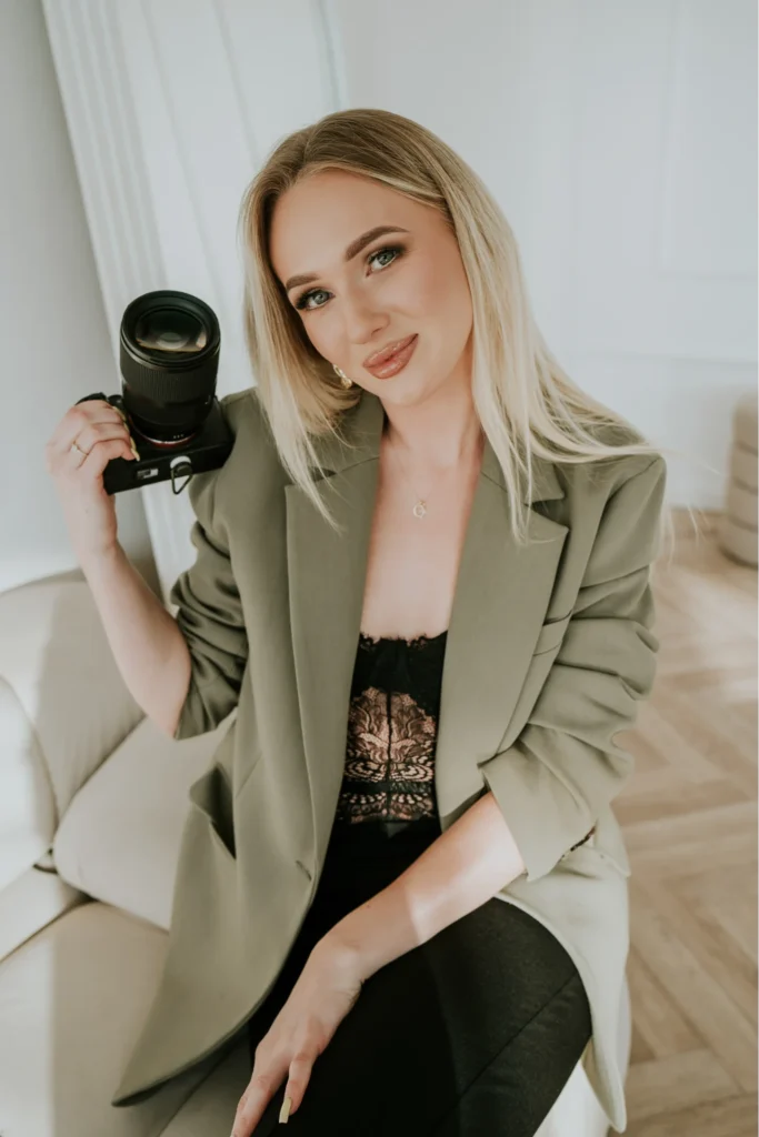 Kobieta w oliwkowej marynarce trzyma aparat fotograficzny z obiektywem siedząc w jasnym wnętrzu